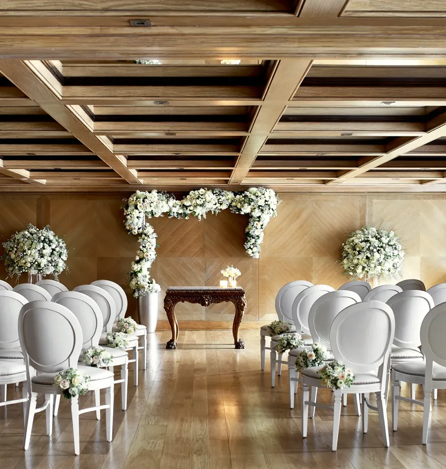 Wedding ceremony room with rows of white chairs facing a floral arch and ornate wooden table under a coffered ceiling.