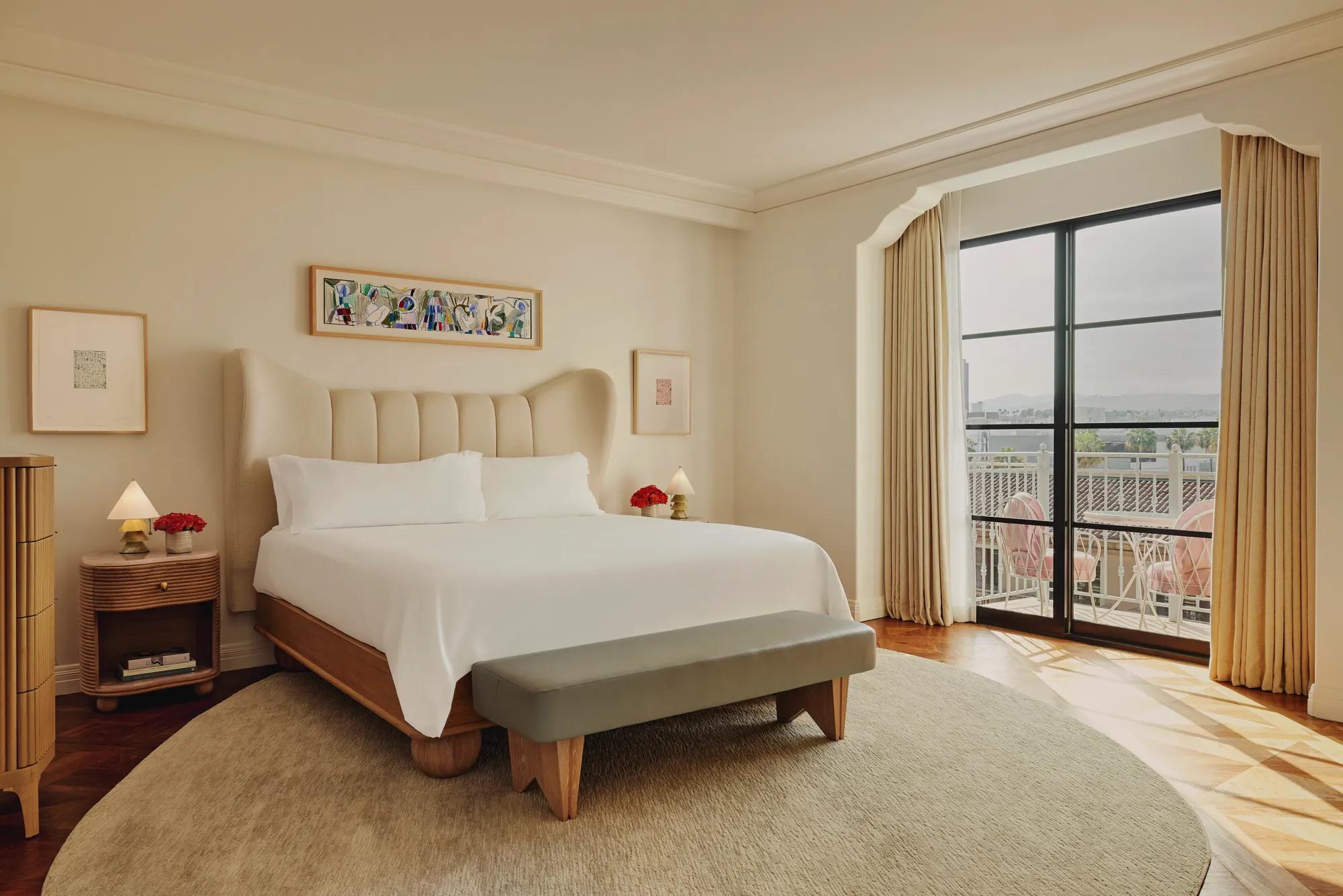 Bright bedroom with a cream upholstered bed, light wood frame, grey bench, and floor-to-ceiling windows leading to a balcony with pink seating.