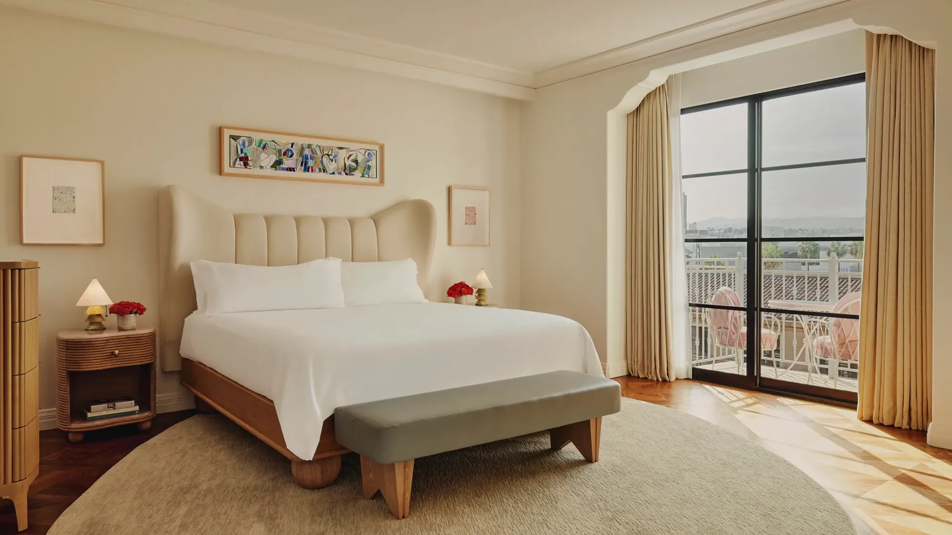 Bright bedroom with a cream upholstered bed, light wood frame, grey bench, and floor-to-ceiling windows leading to a balcony with pink seating.