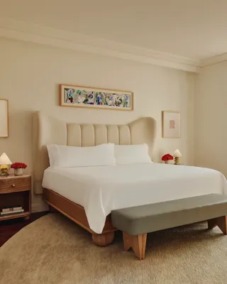 Bright bedroom with a cream upholstered bed, light wood frame, grey bench, and floor-to-ceiling windows leading to a balcony with pink seating.