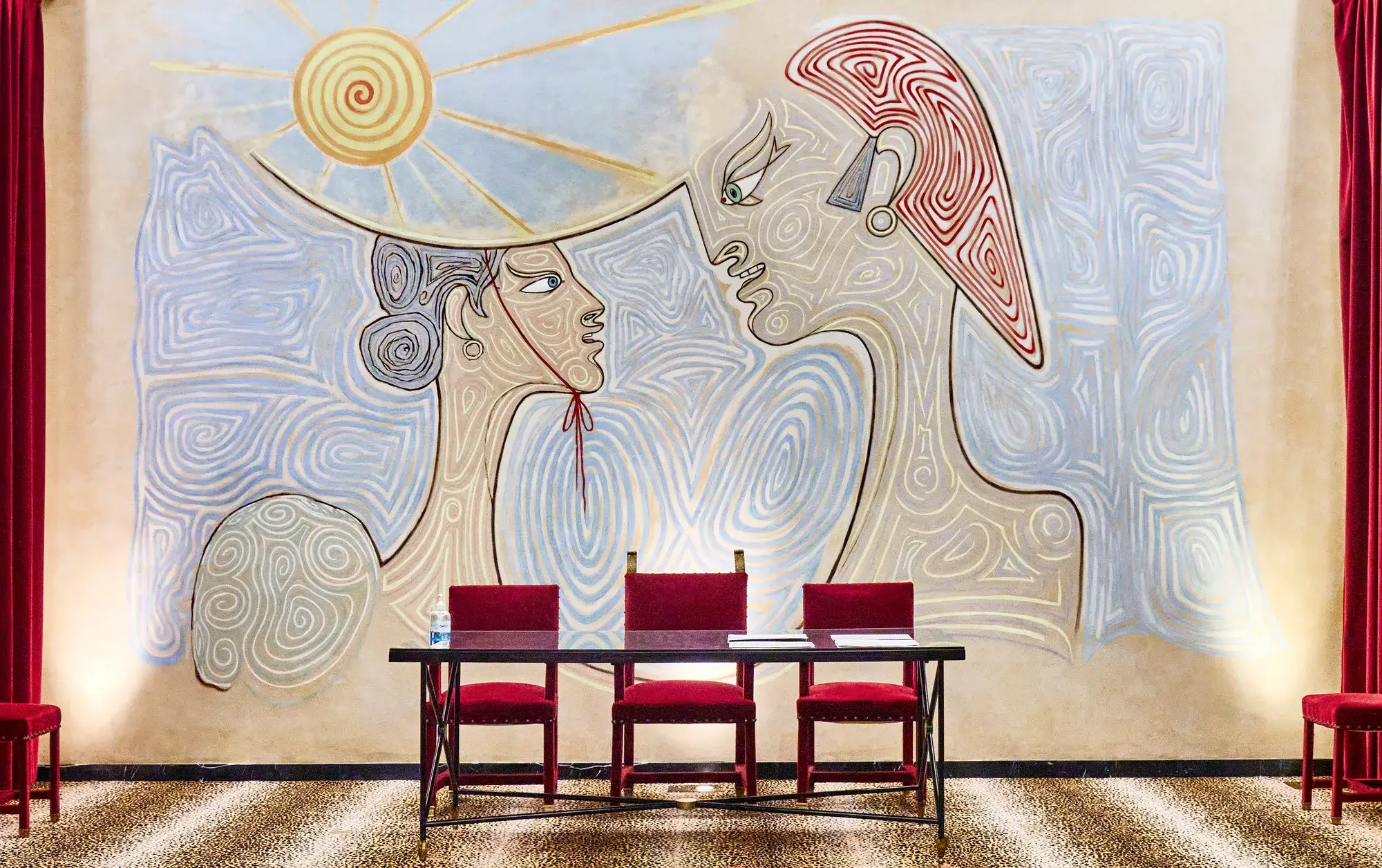 Grande fresque représentant deux figures stylisées se faisant face sous un soleil, au-dessus d’une table et de chaises rouges.