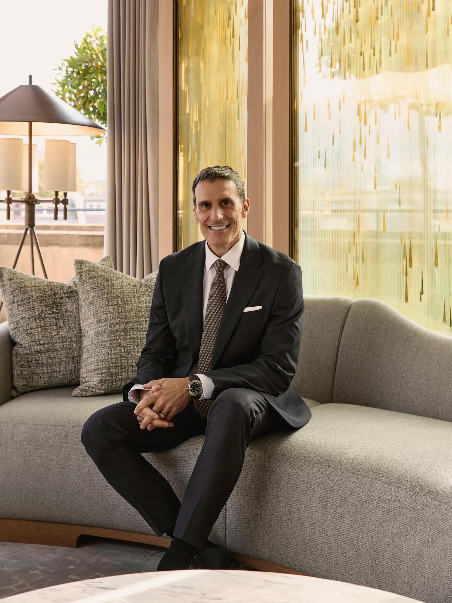 Person in a dark suit seated on a curved sofa in an elegant lounge with floor-to-ceiling windows and warm-toned interiors.