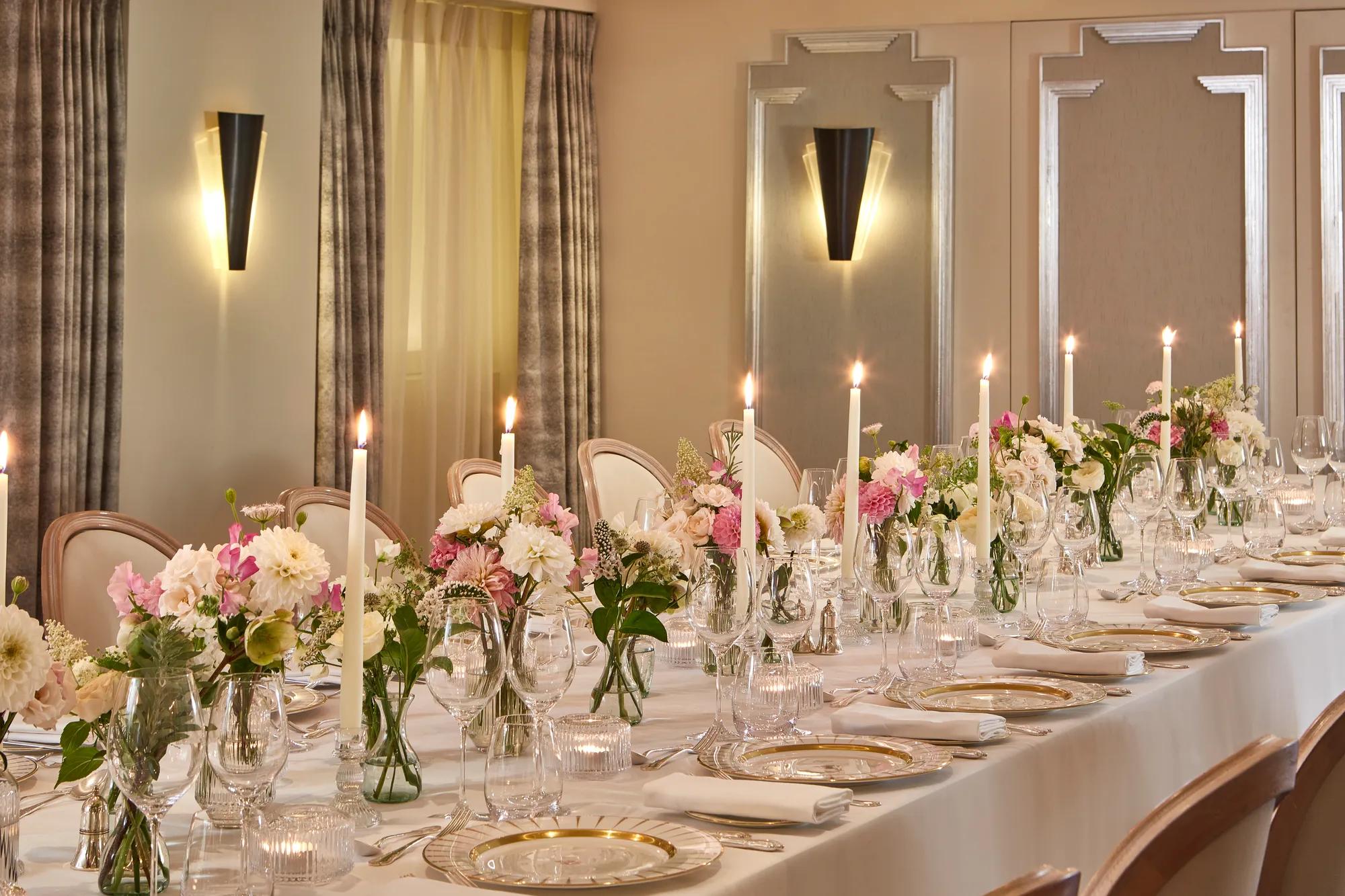 Elegant dining table at Claridge’s set with white linens, crystal glassware, gold-rimmed plates, tall candles, and floral centrepieces in soft pink and white tones.