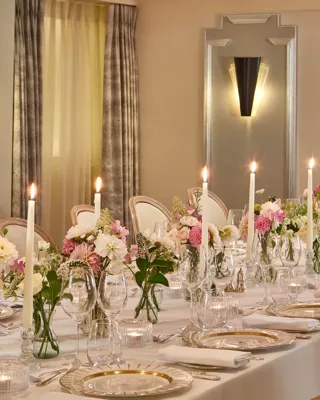 Elegant dining table at Claridge’s set with white linens, crystal glassware, gold-rimmed plates, tall candles, and floral centrepieces in soft pink and white tones.