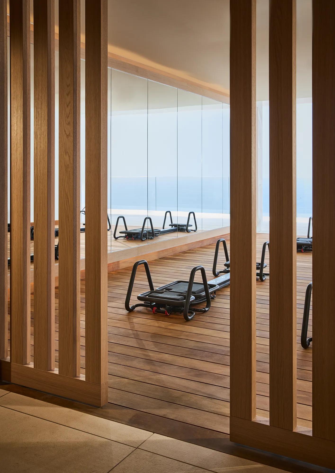 Pilates reformers on a wooden studio deck, viewed through timber screens, with floor-to-ceiling windows overlooking the sea.