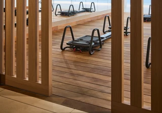 Pilates reformers on a wooden studio deck, viewed through timber screens, with floor-to-ceiling windows overlooking the sea.
