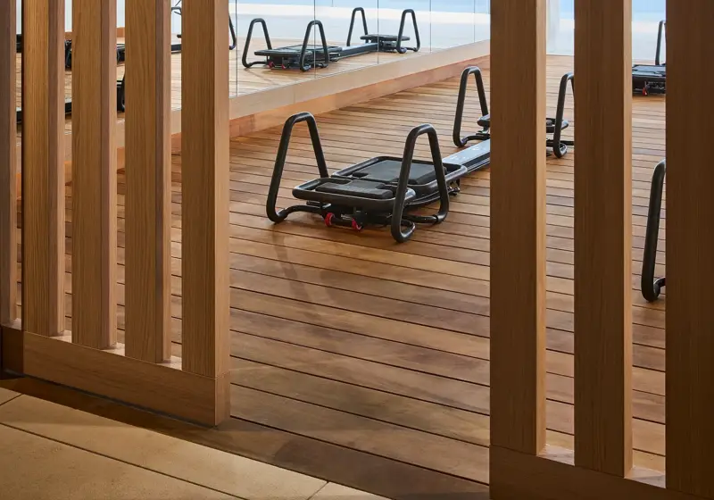 Pilates reformers on a wooden studio deck, viewed through timber screens, with floor-to-ceiling windows overlooking the sea.