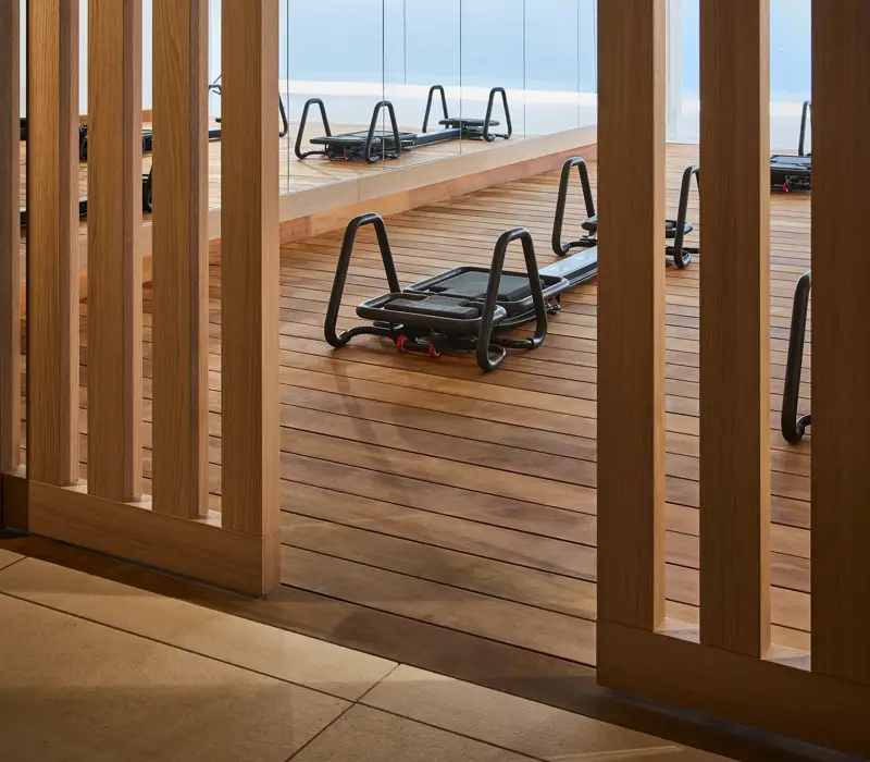 Pilates reformers on a wooden studio deck, viewed through timber screens, with floor-to-ceiling windows overlooking the sea.