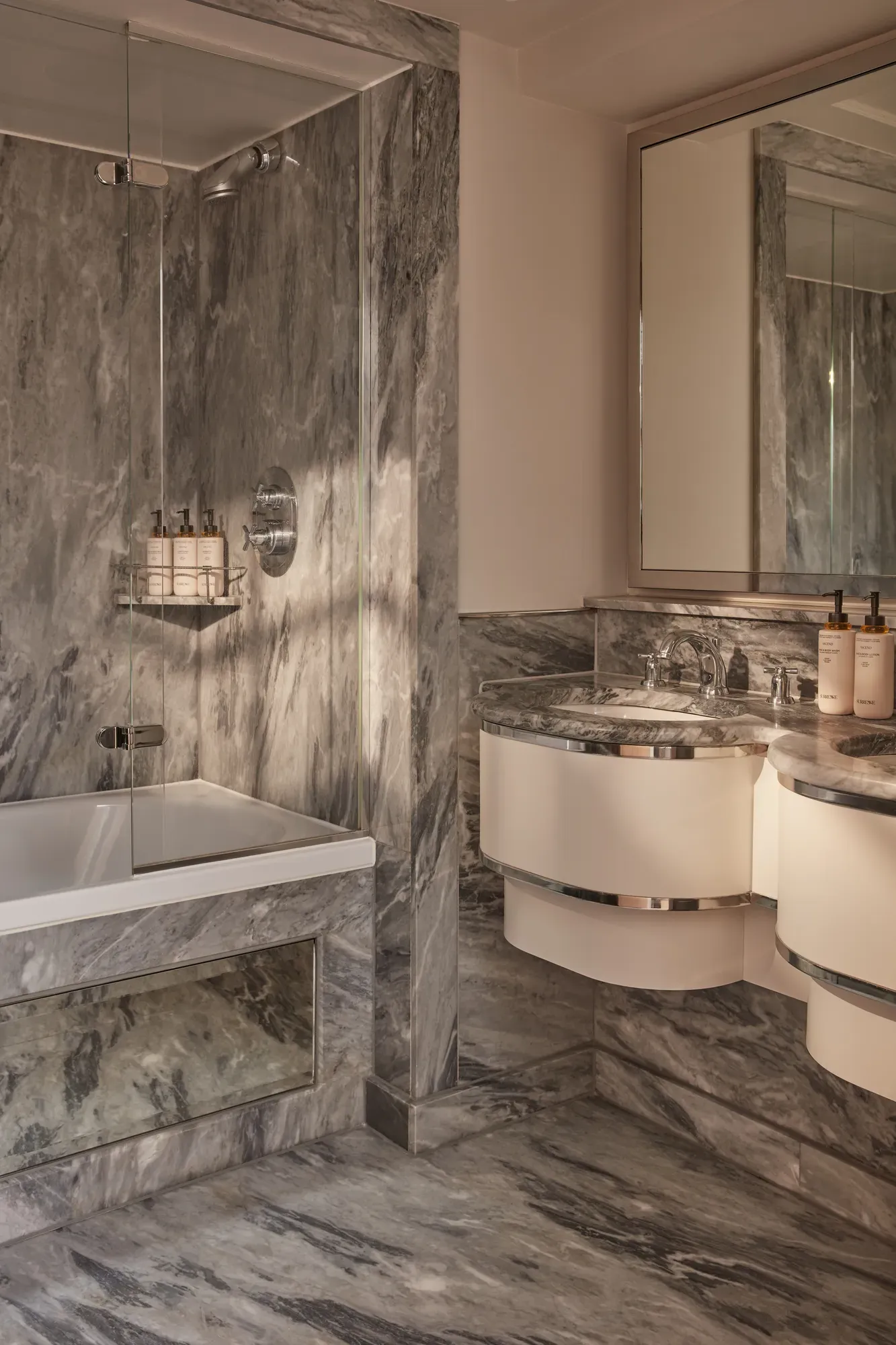 Marble Belgravia Room bathroom with vanity, large mirror, glass-enclosed shower over bathtub, and luxury toiletries.