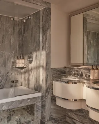 Marble Belgravia Room bathroom with vanity, large mirror, glass-enclosed shower over bathtub, and luxury toiletries.