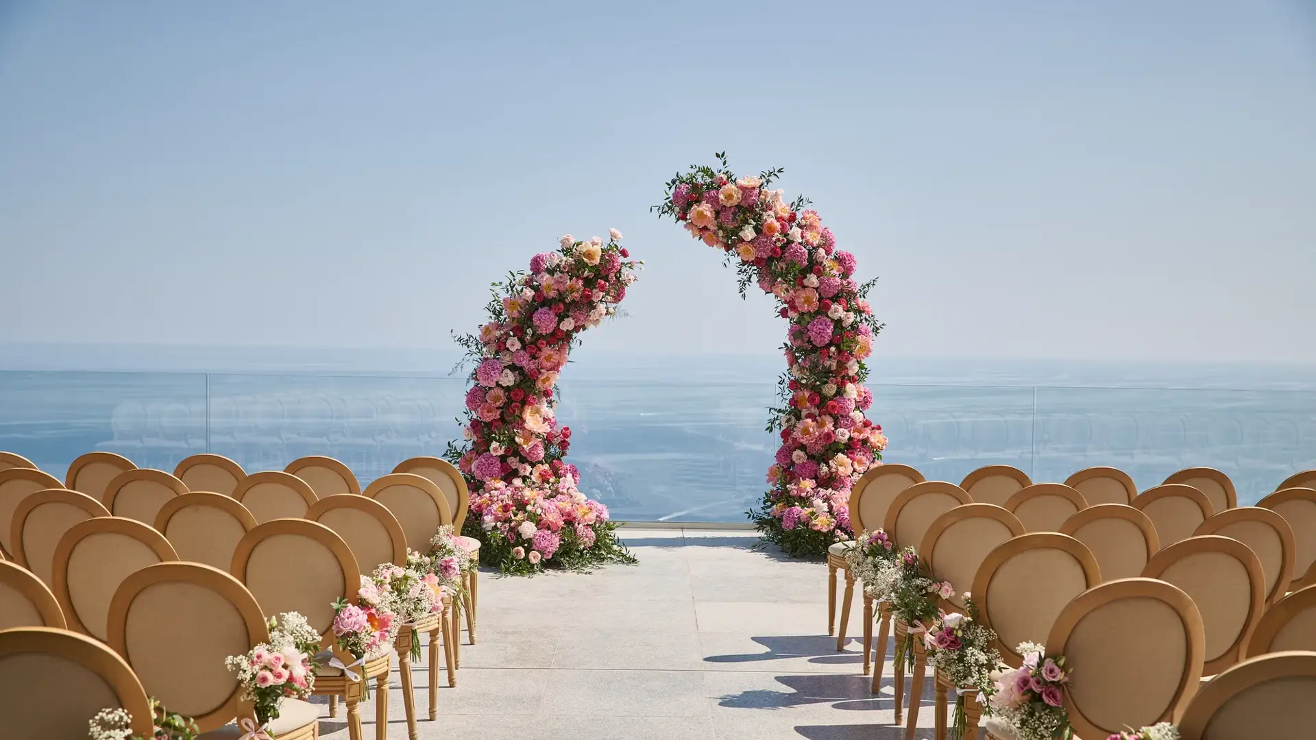 Installation de cérémonie de mariage en plein air avec des rangées de chaises décorées de fleurs roses et blanches, faisant face à deux arches florales incurvées avec en arrière-plan la mer et un ciel dégagé.