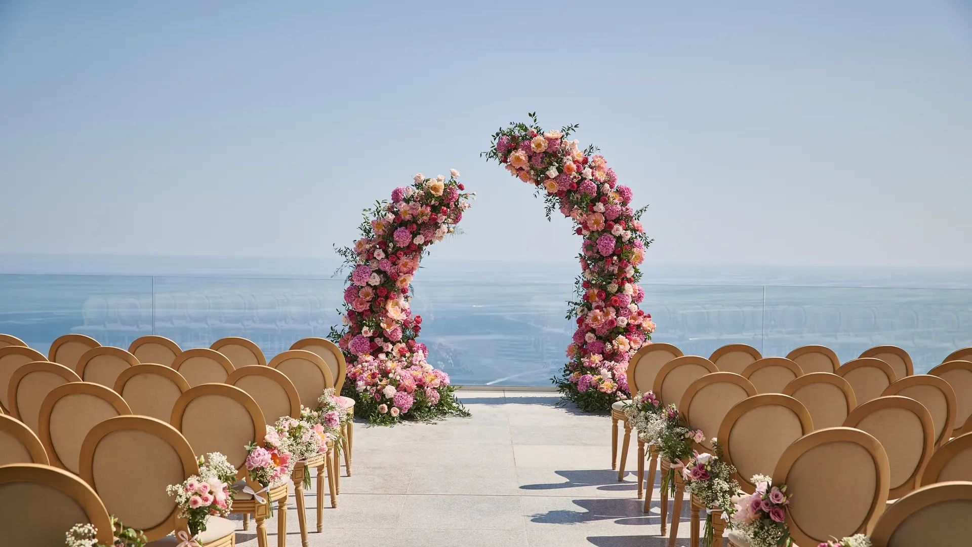 Installation de cérémonie de mariage en plein air avec des rangées de chaises décorées de fleurs roses et blanches, faisant face à deux arches florales incurvées avec en arrière-plan la mer et un ciel dégagé.