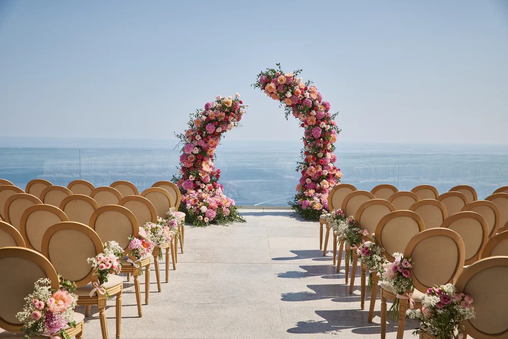 Installation de cérémonie de mariage en plein air avec des rangées de chaises décorées de fleurs roses et blanches, faisant face à deux arches florales incurvées avec en arrière-plan la mer et un ciel dégagé.