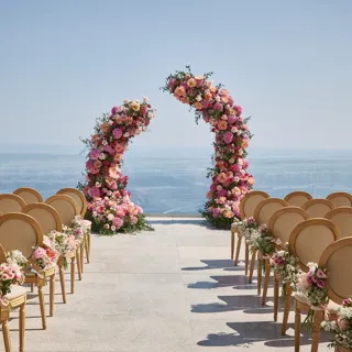 Outdoor wedding ceremony setup with rows of chairs decorated with pink and white flowers, facing two curved floral arches against a backdrop of the sea and clear sky.