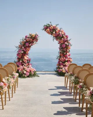 Installation de cérémonie de mariage en plein air avec des rangées de chaises décorées de fleurs roses et blanches, faisant face à deux arches florales incurvées avec en arrière-plan la mer et un ciel dégagé.
