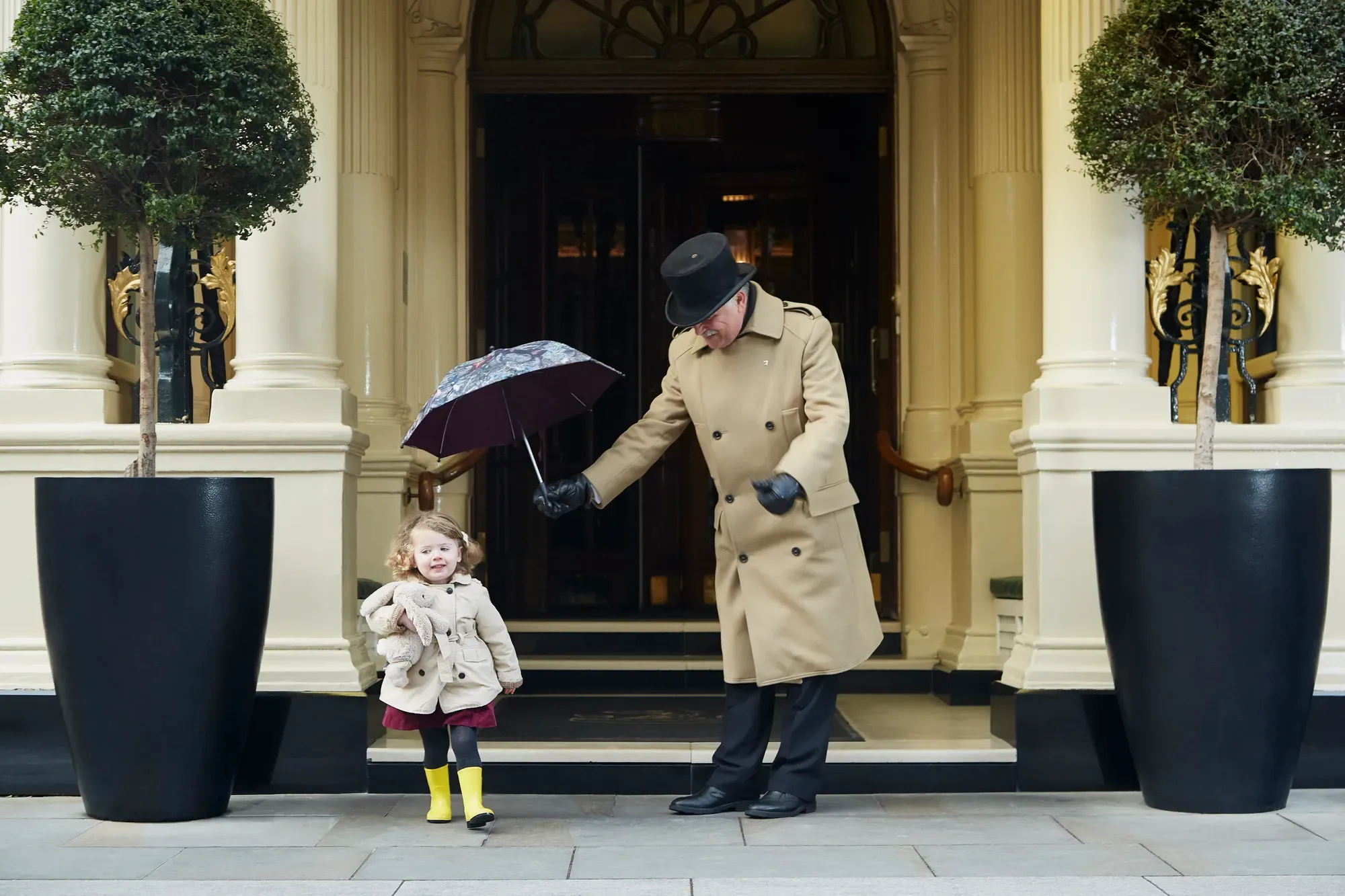A doorman in a tan coat holds an umbrella over a young child in a raincoat and yellow boots outside the hotel’s grand entrance.