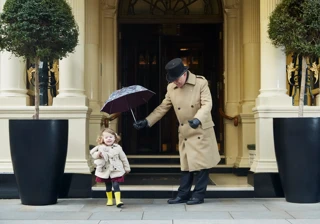 A doorman in a tan coat holds an umbrella over a young child in a raincoat and yellow boots outside the hotel’s grand entrance.