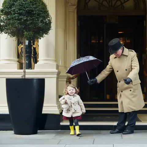 A doorman in a tan coat holds an umbrella over a young child in a raincoat and yellow boots outside the hotel’s grand entrance.