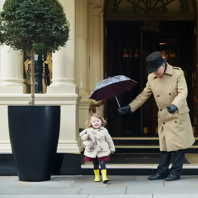 A doorman in a tan coat holds an umbrella over a young child in a raincoat and yellow boots outside the hotel’s grand entrance.