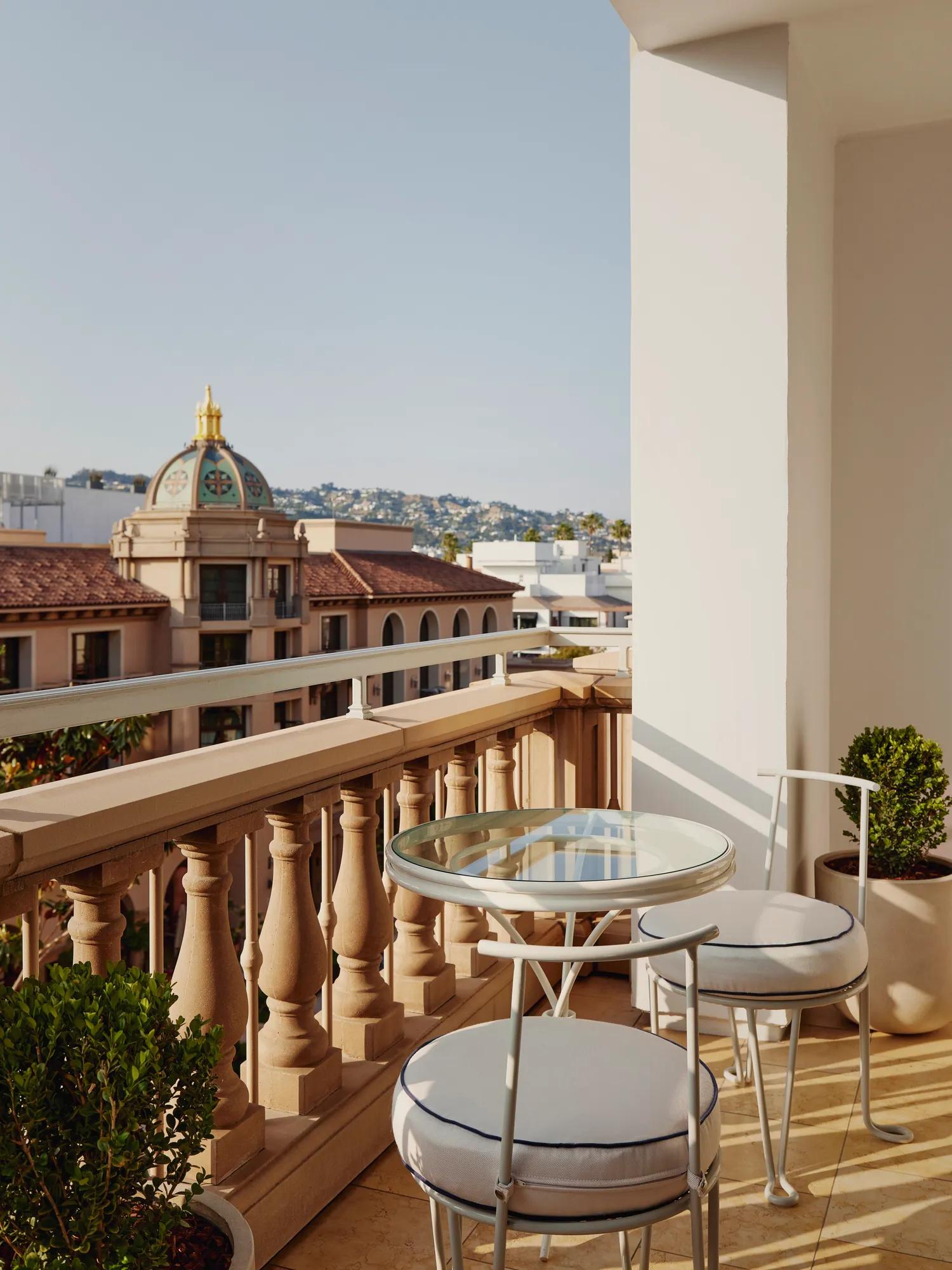 Private hotel balcony with a glass-top bistro table and two cushioned chairs, balustrade, and city and hillside views.