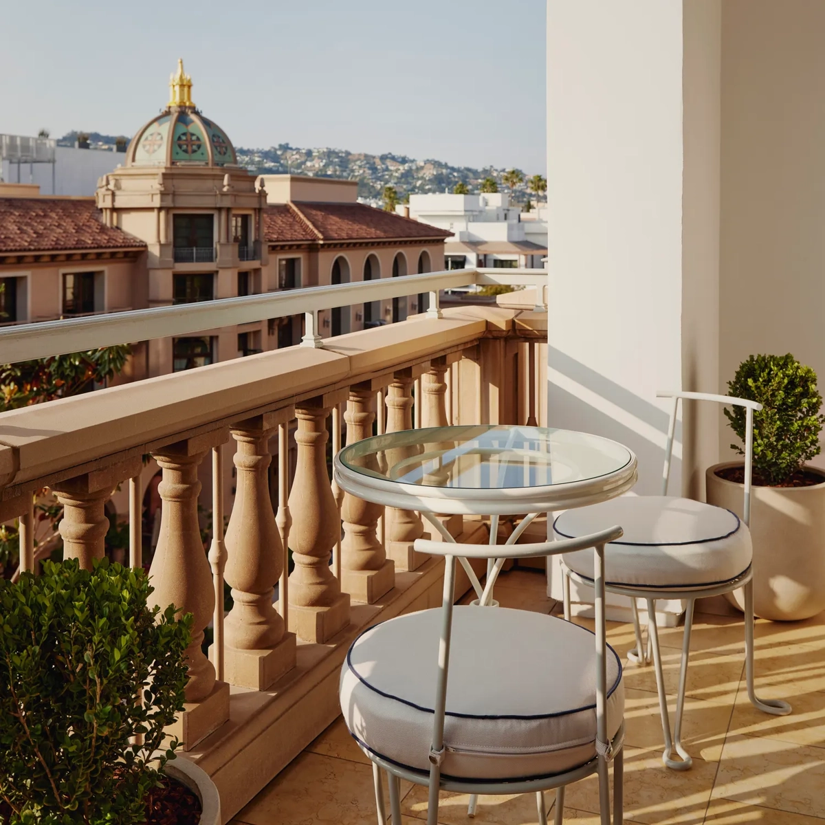 Private hotel balcony with a glass-top bistro table and two cushioned chairs, balustrade, and city and hillside views.