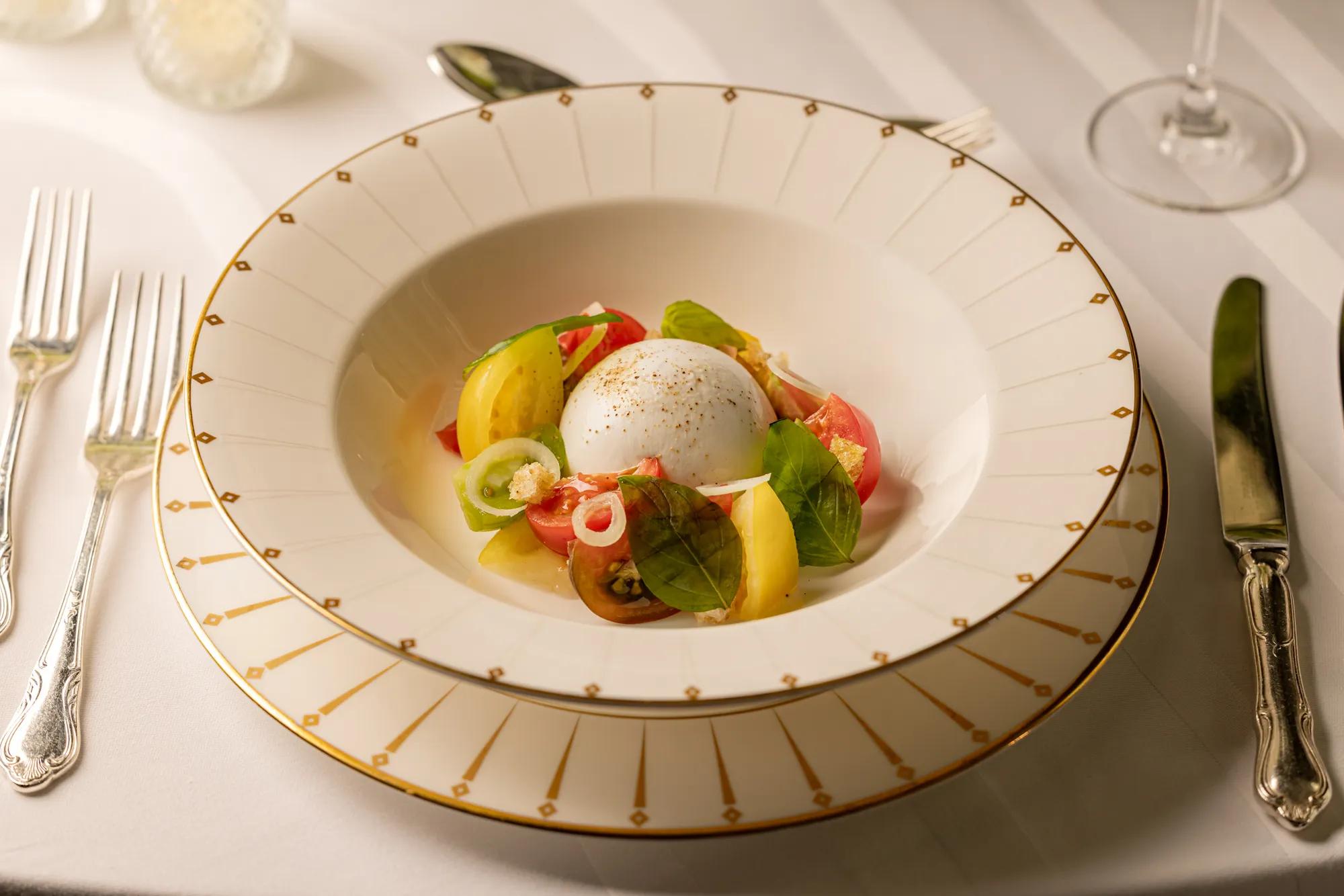 Burrata with heirloom tomatoes, basil leaves and pickled onion served in a white bowl with gold detailing.