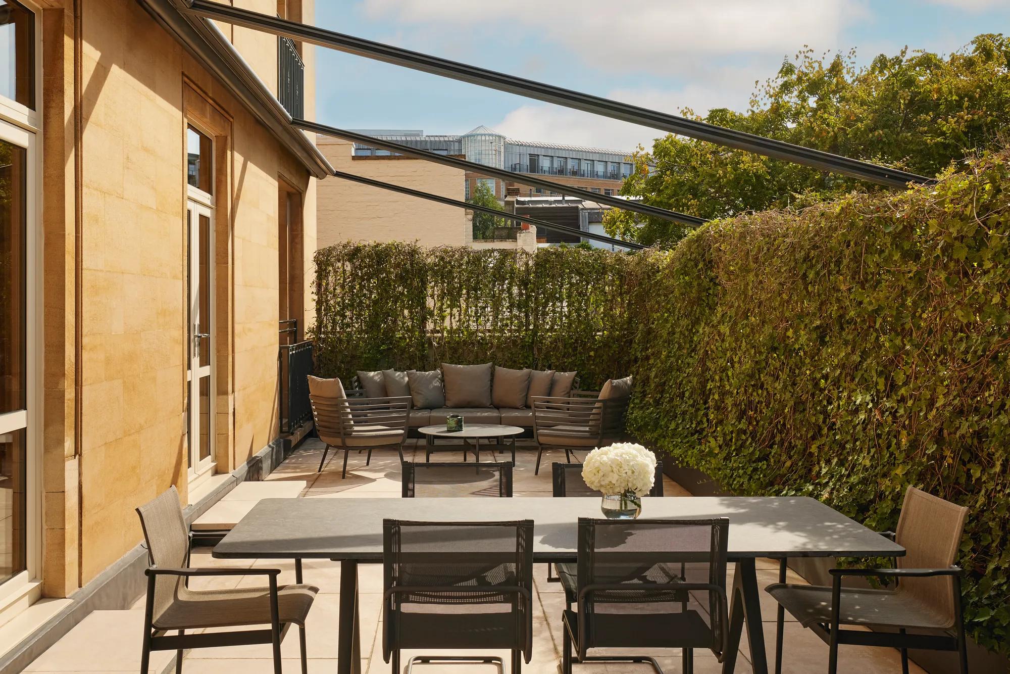 Private terrace with a black dining table, mesh chairs, and a sofa set framed by high green hedges and views of surrounding buildings.