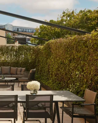 Private terrace with a black dining table, mesh chairs, and a sofa set framed by high green hedges and views of surrounding buildings.