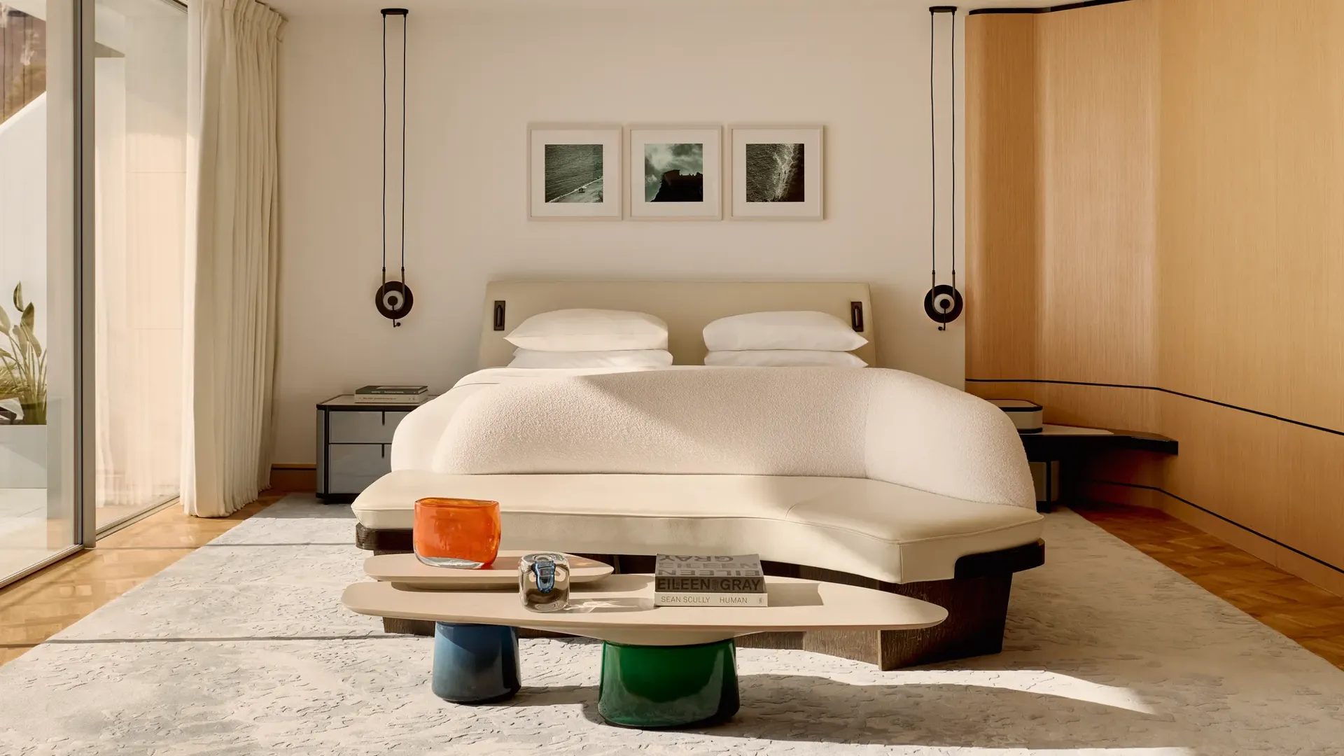 Modern hotel bedroom with large bed, curved bench, and low table, featuring wooden panelling and glass doors opening to a terrace.