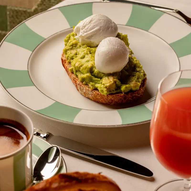 Avocado toast topped with two poached eggs on a striped plate, served with coffee and juice.