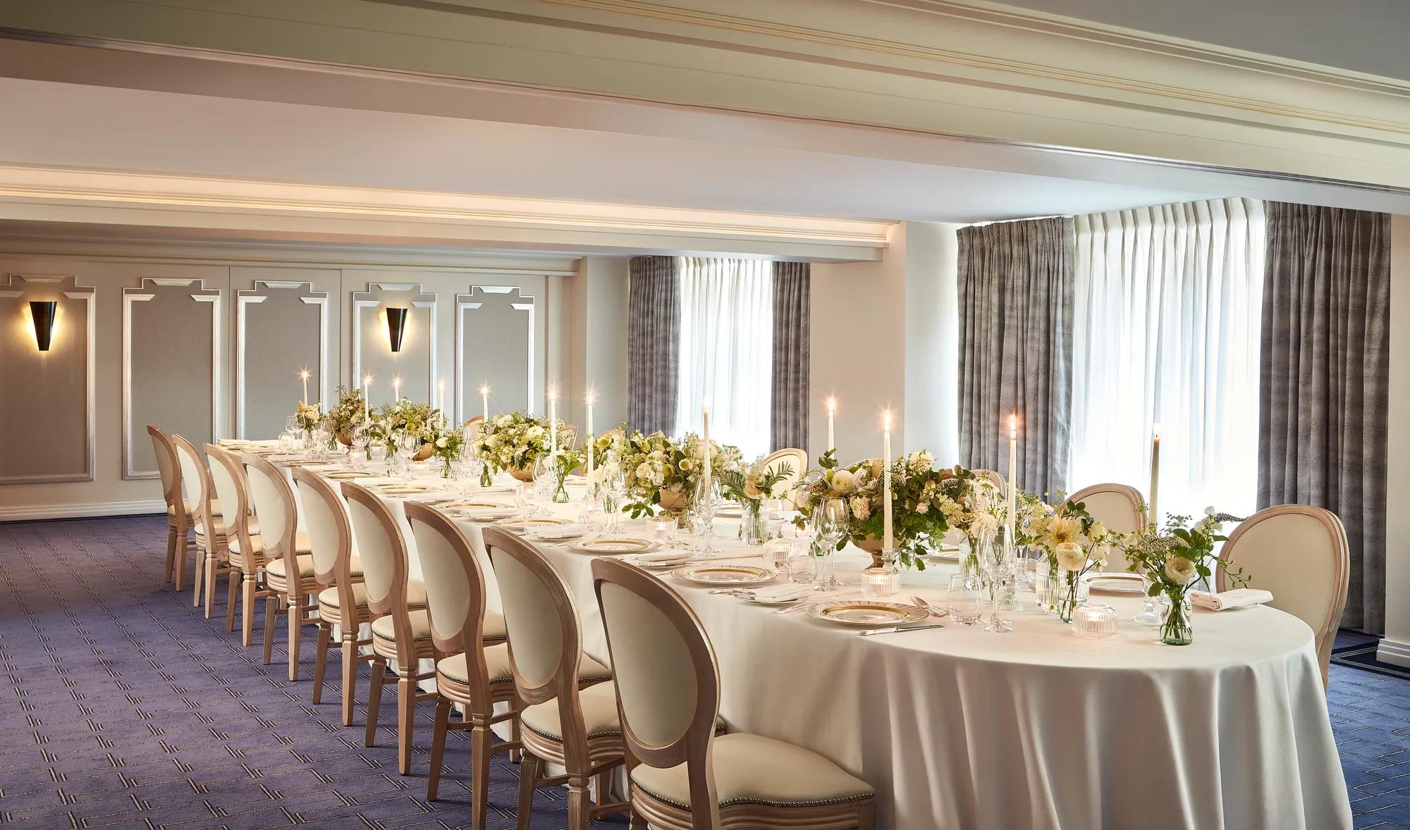 The St James Room at Claridge’s set for a private dinner with an elegant U-shaped table arrangement, white linen, floral centrepieces, and candlelight by the windows.