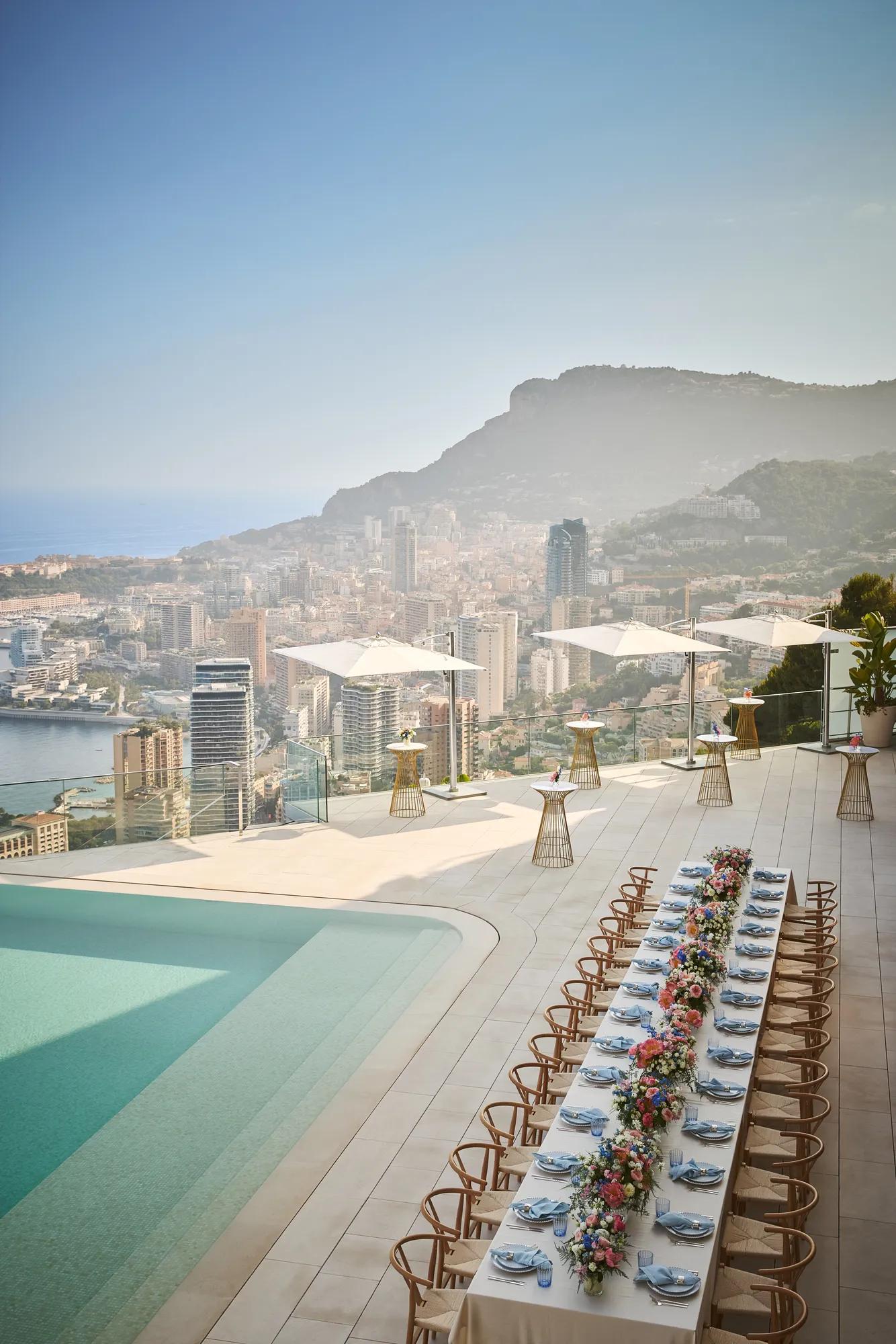 Elegant banquet table set with flowers beside an infinity pool, overlooking a city skyline and mountains under a clear sky.