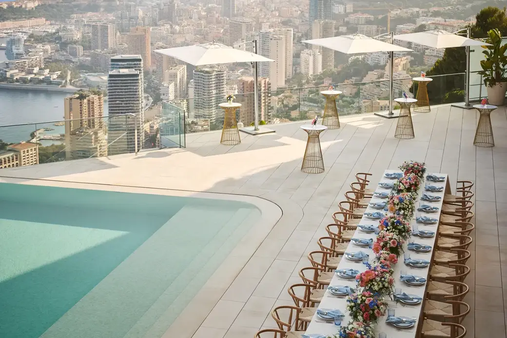 Table de banquet élégante décorée de fleurs près d’une piscine à débordement, avec vue sur la ville et les montagnes sous un ciel dégagé.