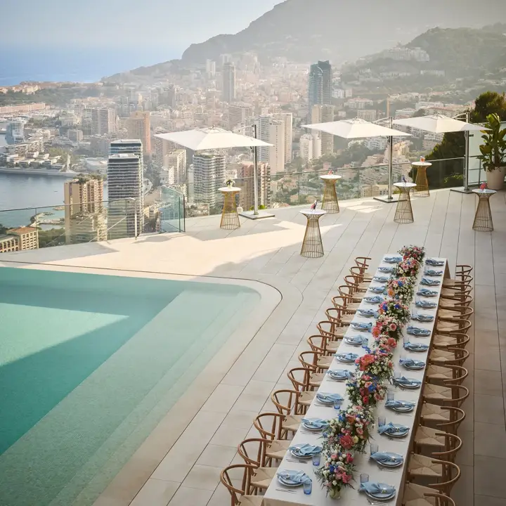 Elegant banquet table set with flowers beside an infinity pool, overlooking a city skyline and mountains under a clear sky.