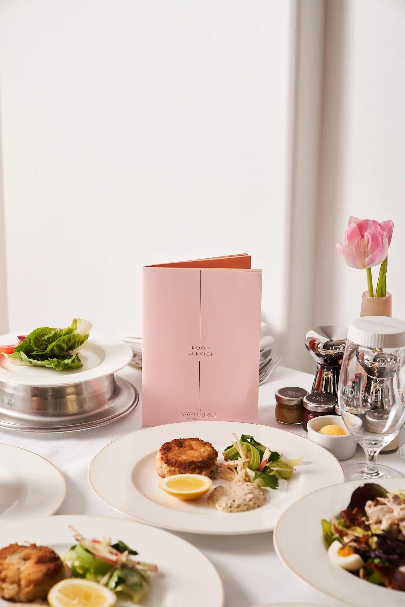 Room service table with crab cakes, salads, and pink menu at The Maybourne Beverly Hills.