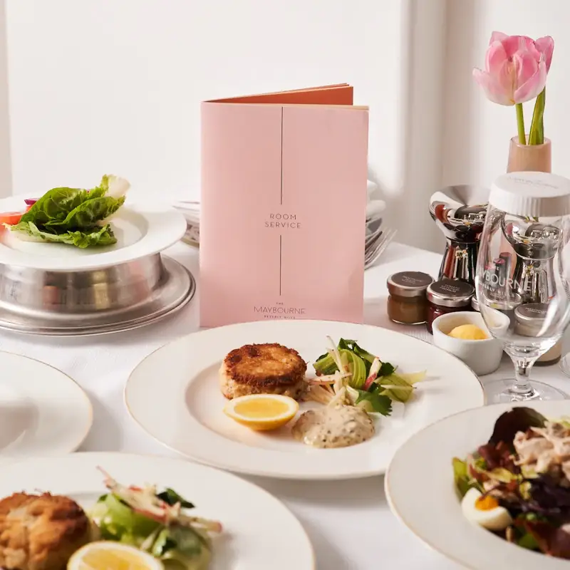 Room service table with crab cakes, salads, and pink menu at The Maybourne Beverly Hills.