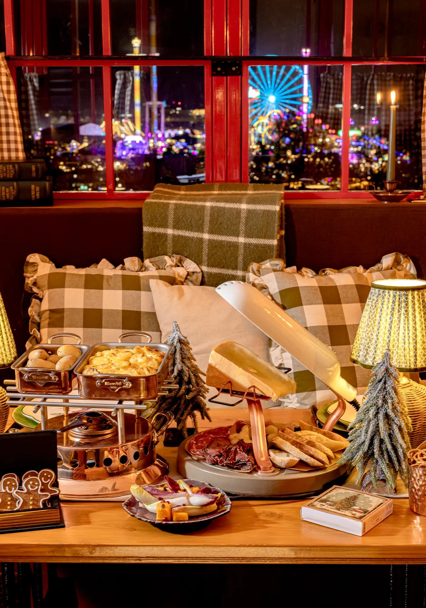 Cosy festive dining scene with raclette, charcuterie and checked cushions, overlooking Christmas lights and a Ferris wheel.
