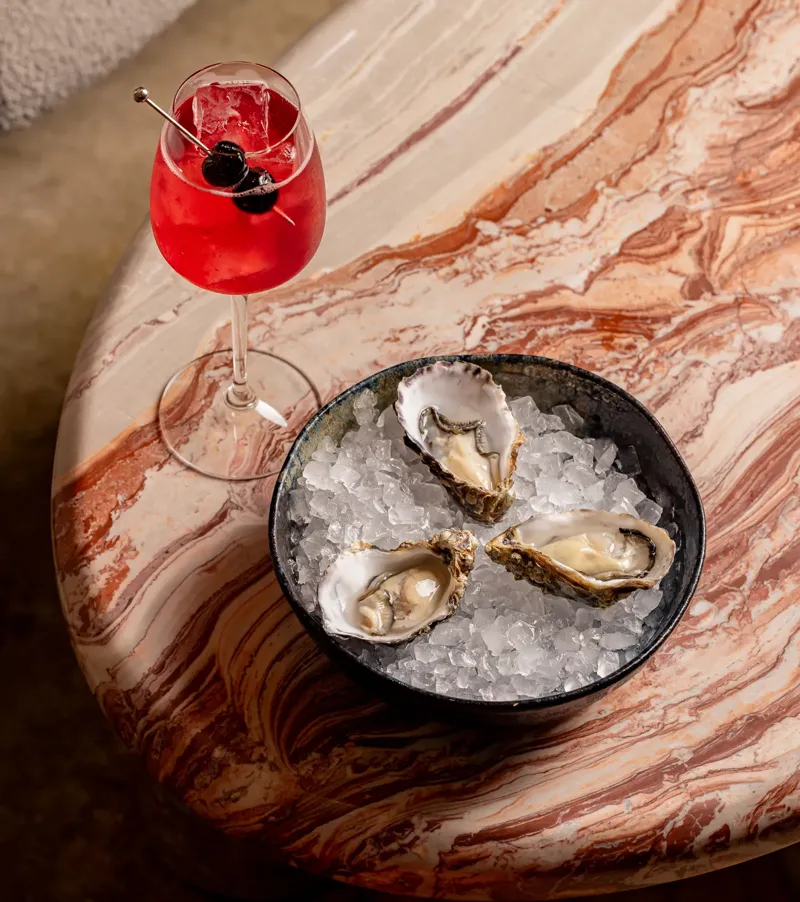 Red cocktail with cherries and a bowl of oysters on ice, presented on a polished marble table.