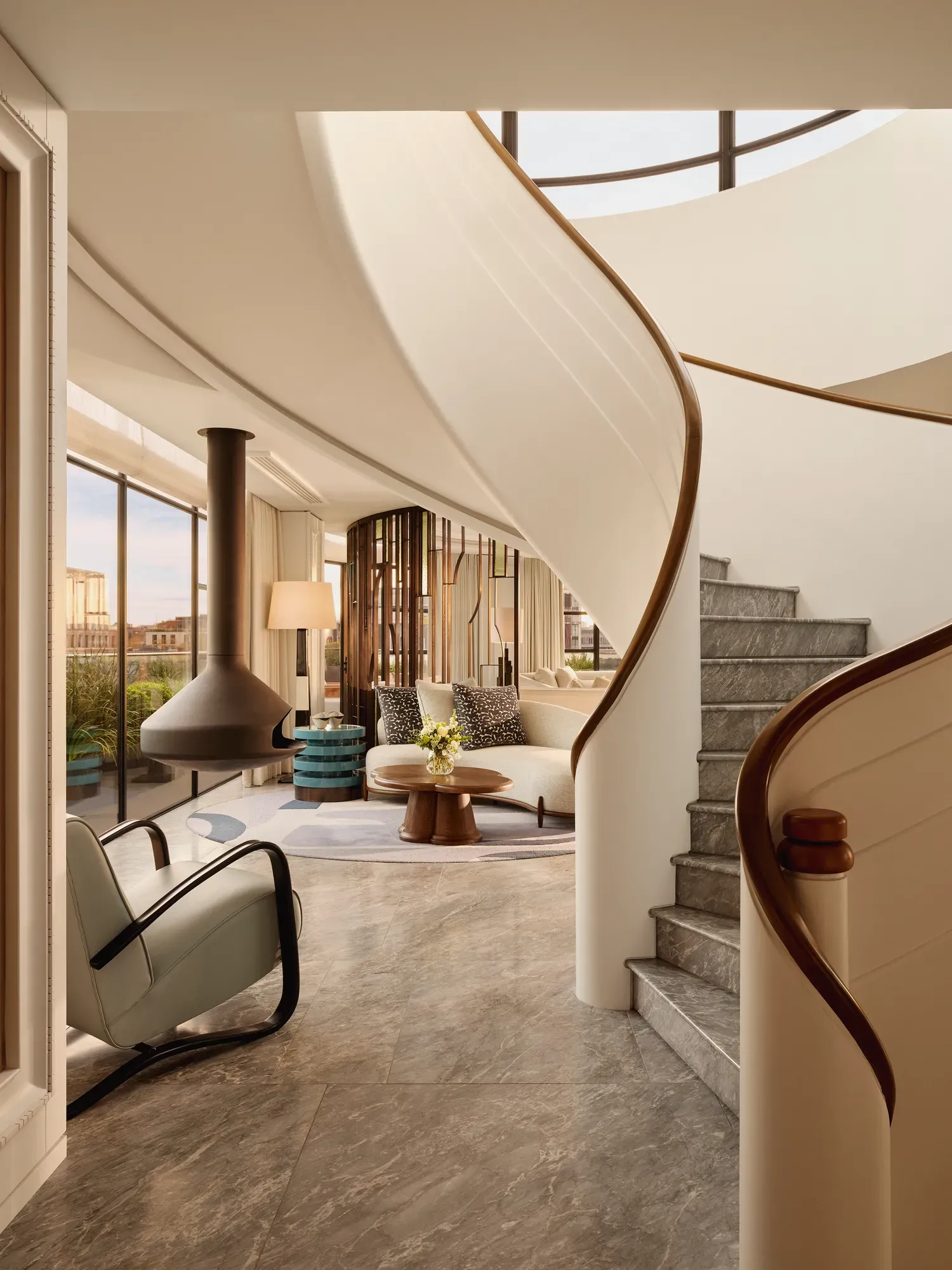 Curved staircase leading to a bright living area with floor-to-ceiling windows, modern furniture, and a suspended fireplace at Claridge’s