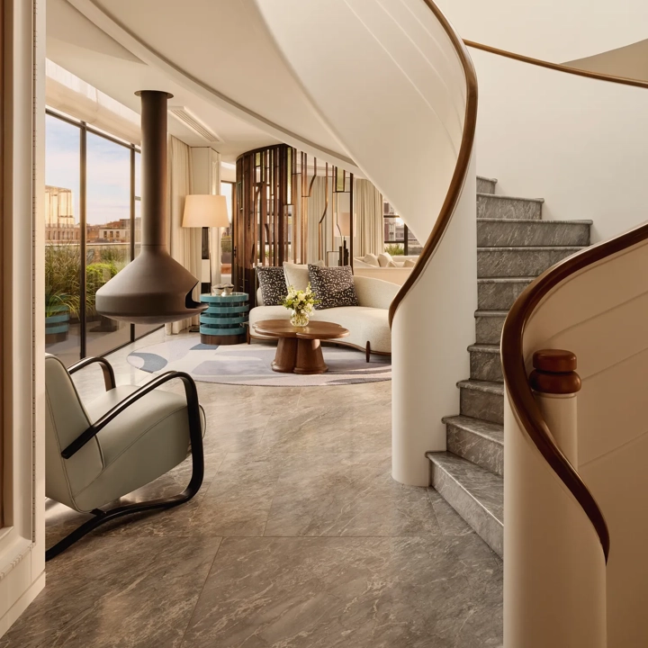 Curved staircase leading to a bright living area with floor-to-ceiling windows, modern furniture, and a suspended fireplace at Claridge’s