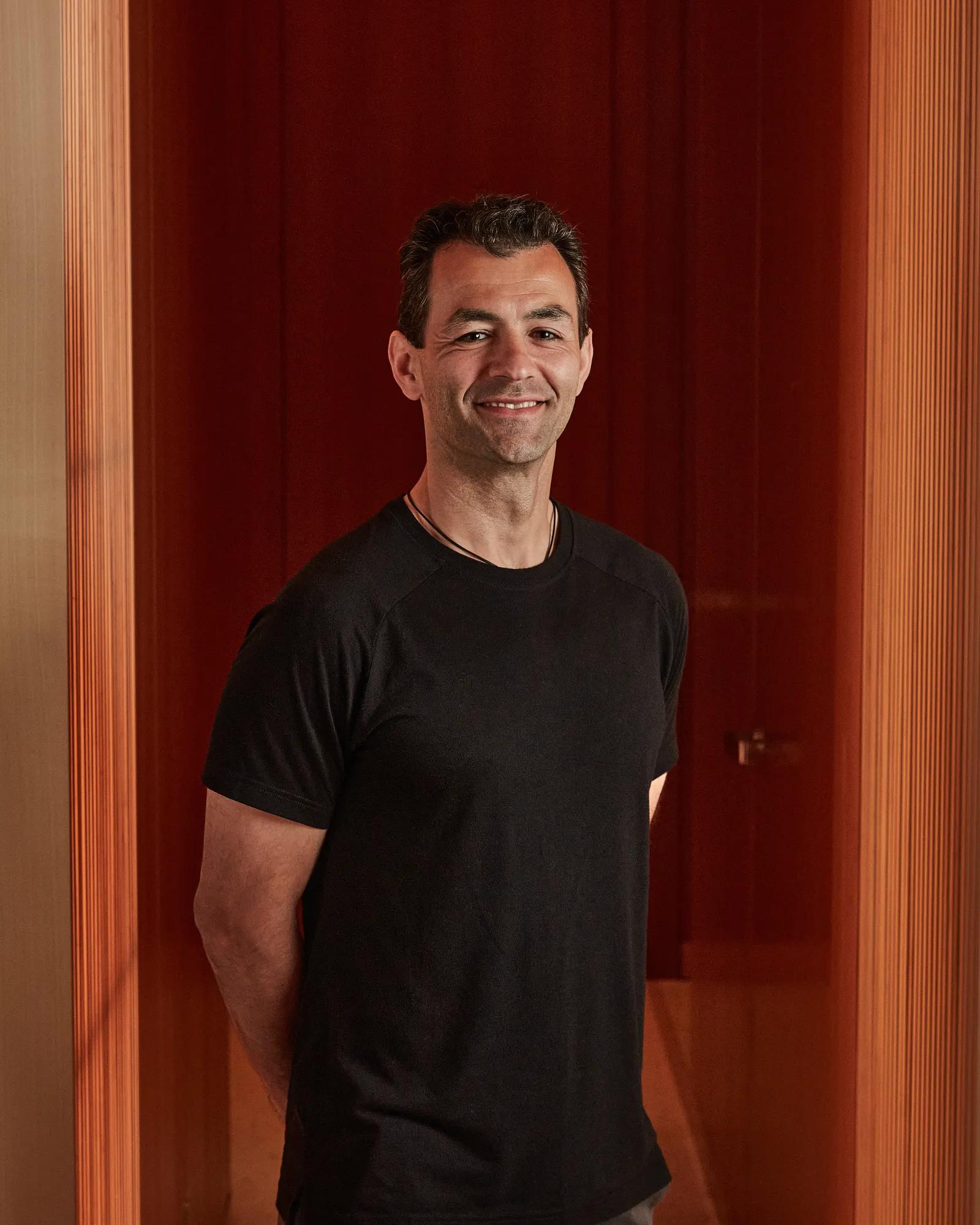 Man in black T-shirt smiling against red paneled background.