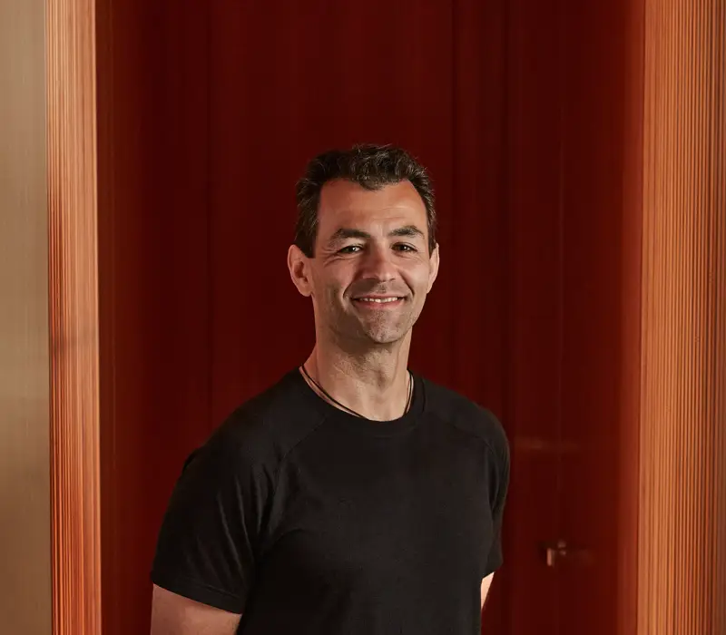 Man in black T-shirt smiling against red paneled background.