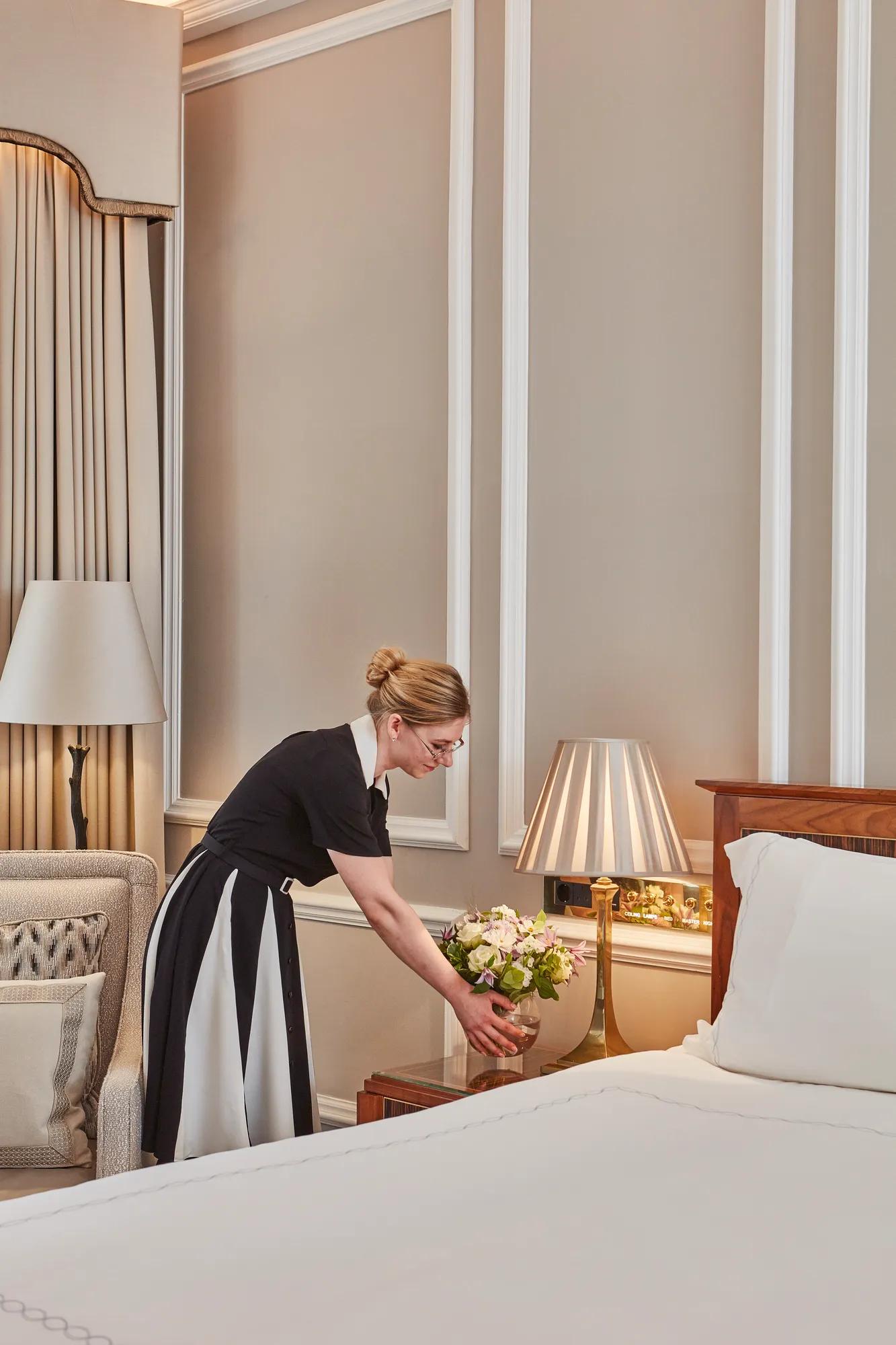 Housekeeper arranging a vase of fresh flowers beside a neatly made luxury hotel bed.
