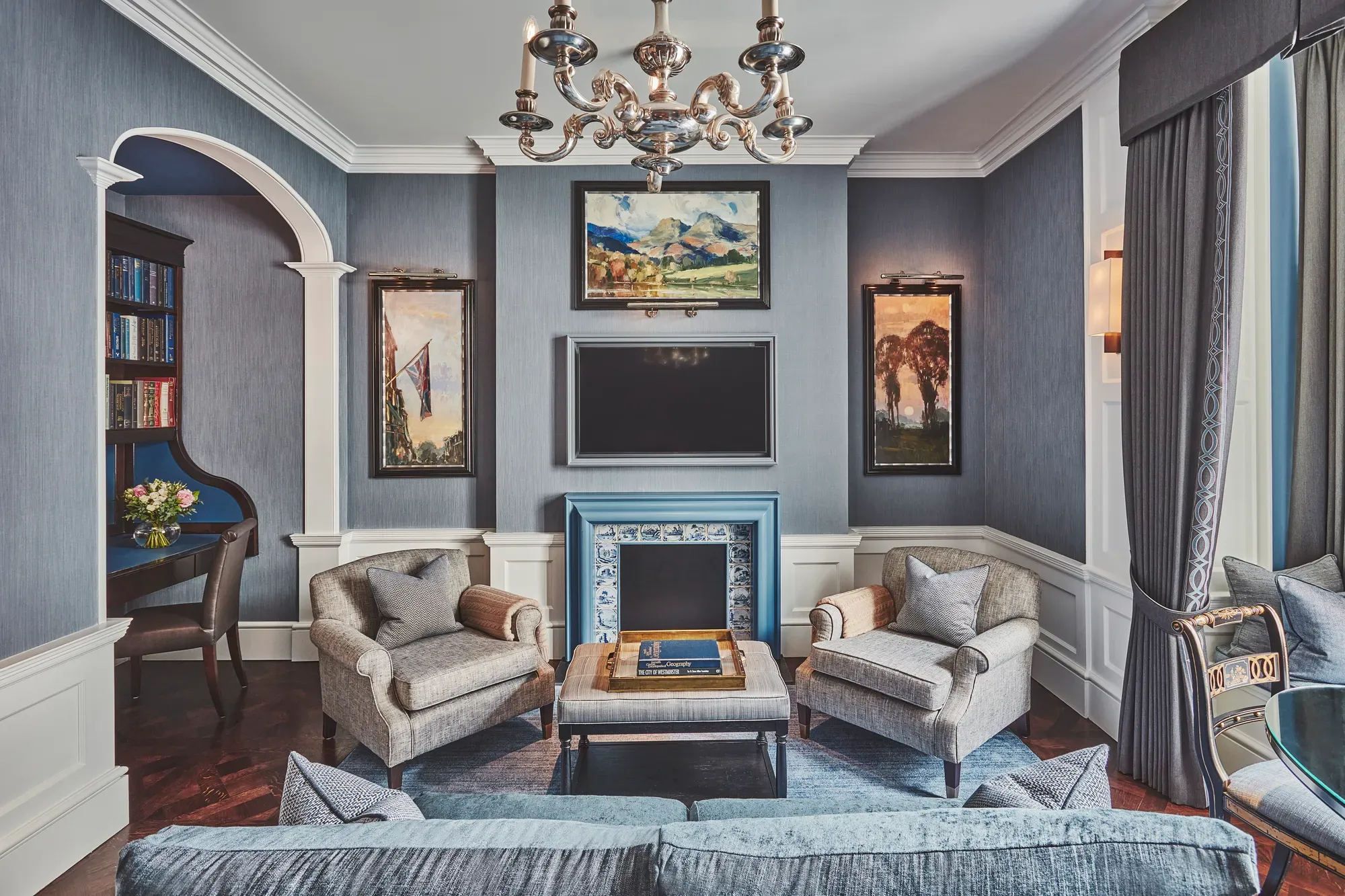 Refined sitting room with grey seating, blue-tiled fireplace, chandelier, and built-in study nook.