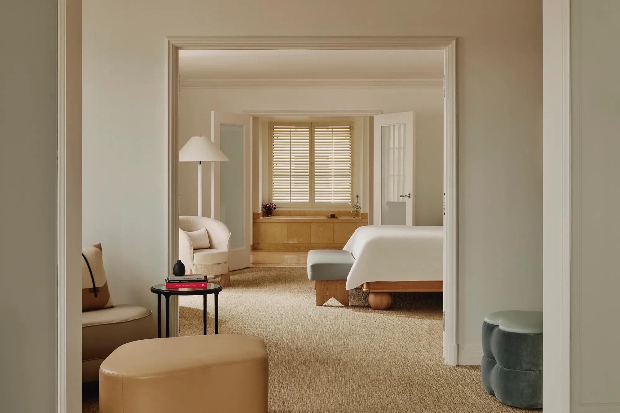 Enfilade view from lounge through bedroom to bathing alcove; shuttered window over tub; neutral upholstery, ottomans, and a small side table stacked with books.