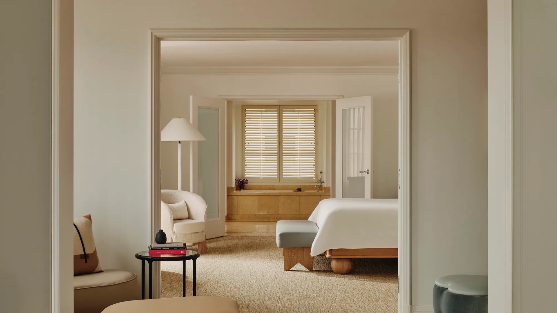 Enfilade view from lounge through bedroom to bathing alcove; shuttered window over tub; neutral upholstery, ottomans, and a small side table stacked with books.