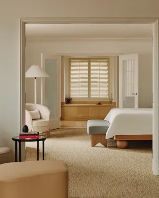 Enfilade view from lounge through bedroom to bathing alcove; shuttered window over tub; neutral upholstery, ottomans, and a small side table stacked with books.
