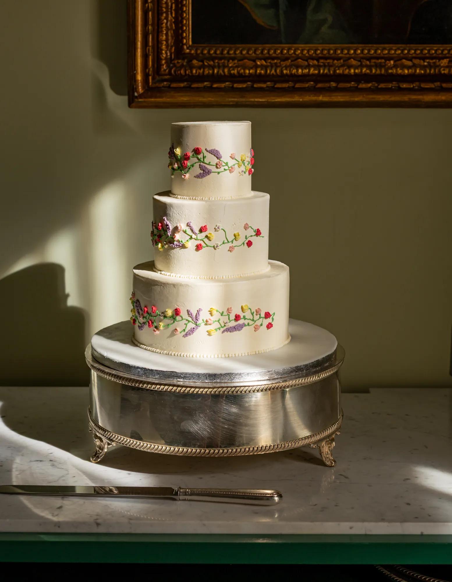 Three-tier white cake with delicate floral decoration displayed on a silver stand in soft natural light