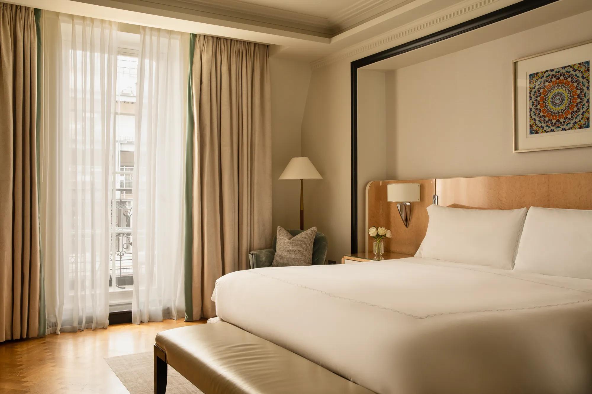 Spacious bedroom with a king-size bed, floor-to-ceiling windows, armchair, bedside table and framed artwork in soft neutral tones.