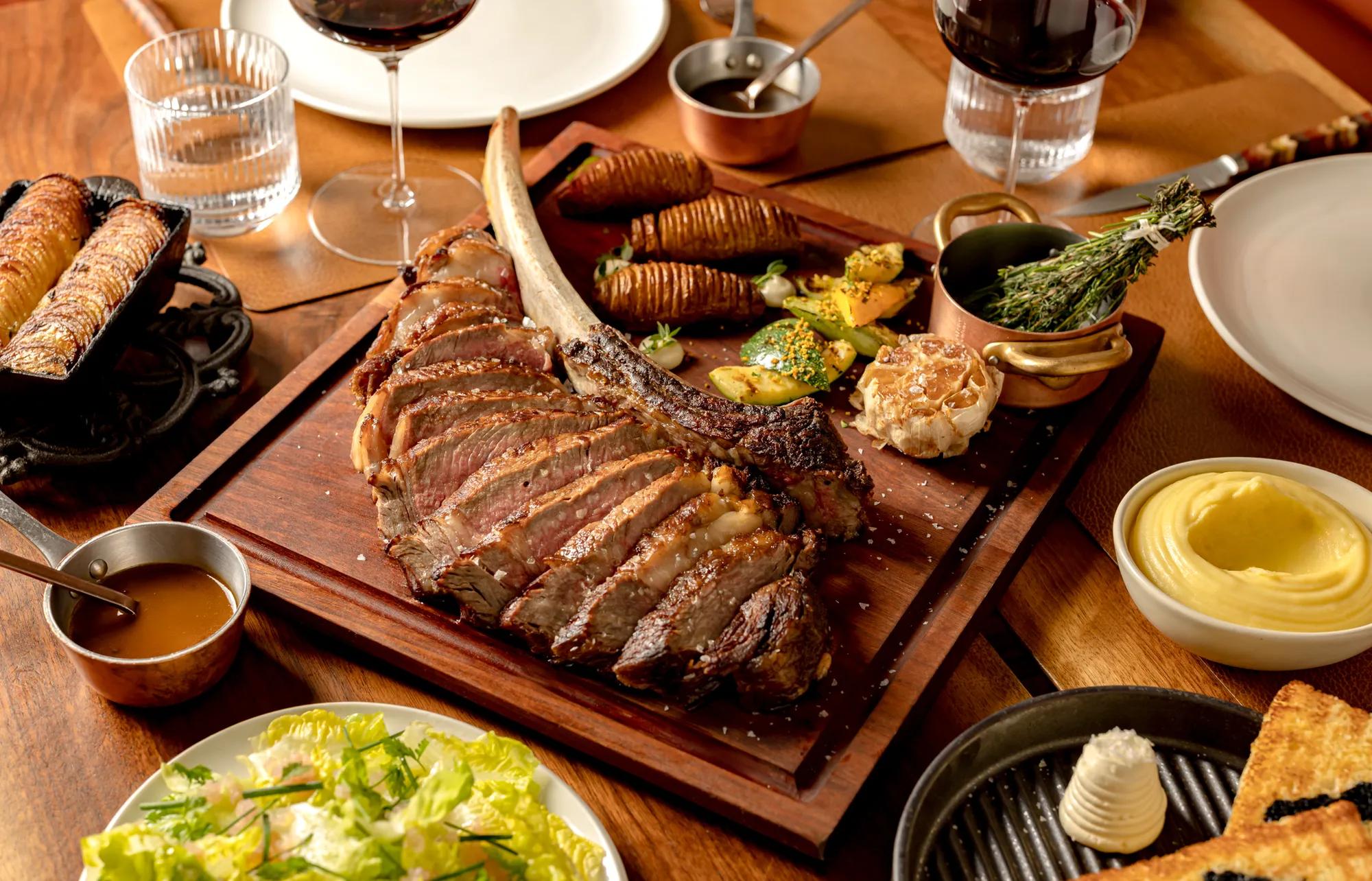 Tomahawk steak served on a wooden board with roasted potatoes and side dishes on a dining table.